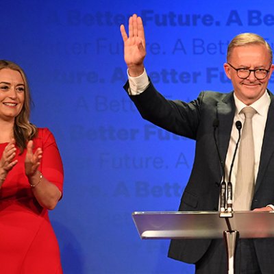 A man at a lecturn waving and a woman in a red dress watching on.